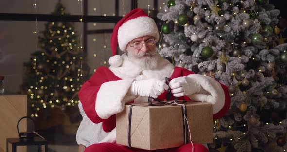 Handsome Elderly Santa Claus Concentrating on Packing a Gift Box Trying to Tie a Bow with a Satin