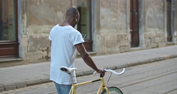 Young African American Man with Earphones Using His Smartphone and Pushing the Stylish Modern Bike