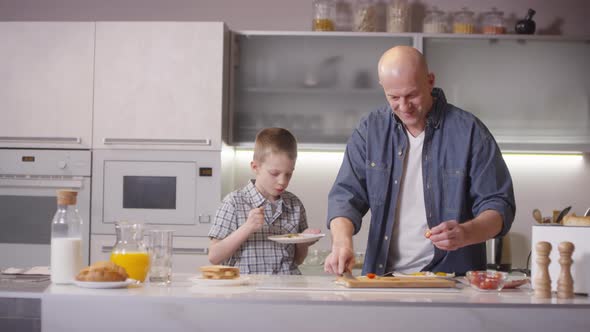 Boy Eating Fried Eggs in Kitchen while his Father Cooking