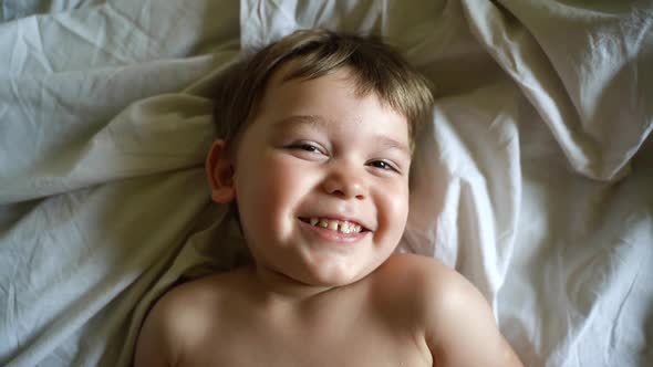 Close-up Portrait of Face of Cheerful Little Boy Lying in Bed on White Sheet and Looks at Camera