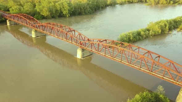 Aerial view of a narrow bridge over muddy wide river in green rural area.