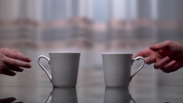 A man and a woman take white ceramic mugs from the kitchen table, against a dark window