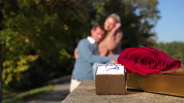 Closeup on a Wedding Ring, a Present and a Red Rose in a Park - a Happy Couple Hugs in Background