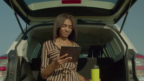 Pretty African Woman Tourist in Car Trunk Networking on Tablet Pc in Nature