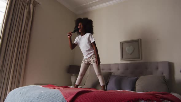 African american girl with hairbrush singing while standing on the bed at home