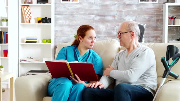 Social Worker Reading To an Old and Disabled Man Sitting on a Sofa in Nursing Home