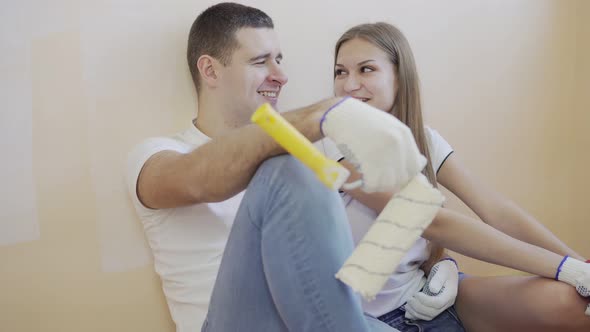 Young Couple Taking a Break From Renovating and Sitting on the Floor with Paint Rollers and Brushes