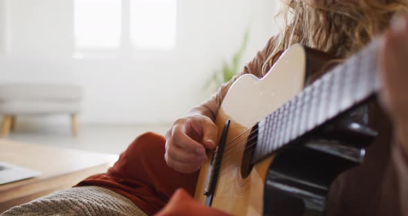 Midsection of caucasian woman playing acoustic guitar, sitting in sunny cottage living room