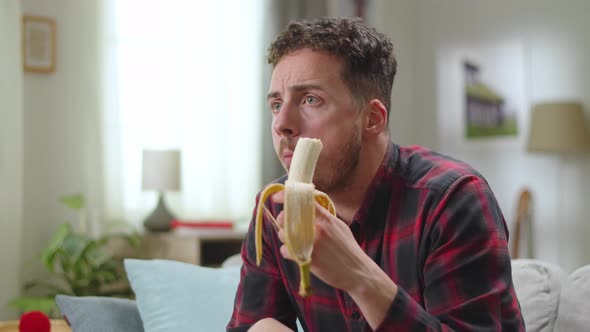 Young Handsome Man Eats Banana and Watching Tv at Home in the Living Room