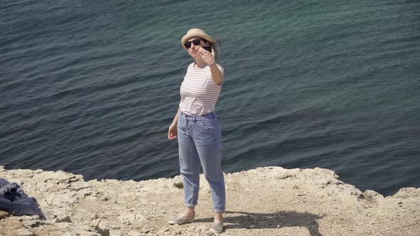 A Woman in a Straw Hat and Sunglasses Takes a Photo By the Sea