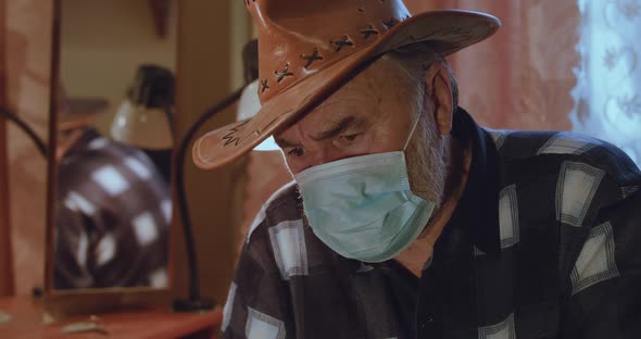 Portrait of Ill Senior Man in Hat and Medical Mask Looks at Camera with Sadness