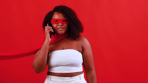 Latin Hispanic Woman in Red Eye Mask and Long Superhero Cloak in Studio Background Talking By Phone