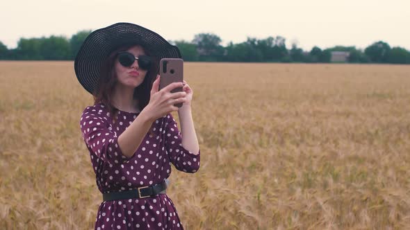 Attractive Fun Hippie Woman in the Wheat Field at Sunset Making a Selfie with Smartphone