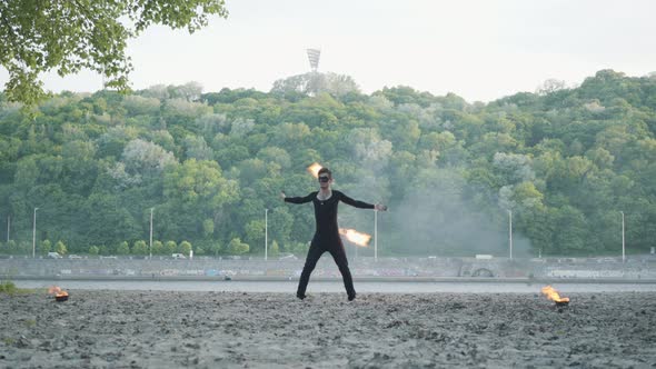 Young Slim Man in Black Clothes and Mask Performing a Show with Flame Standing on Riverbank