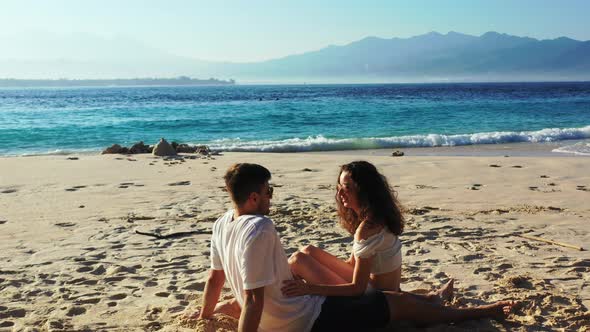 Young couple in love sitting on white sand of exotic beach with blue sea background having a convers