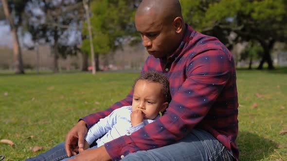 Father Sitting on Grass in Park, Holding Son on Knees, Hugging