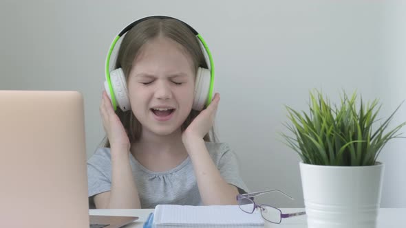Joyful Kid Girl Wearing Headphones Sitting at Desk at Home Opposite Computer Enjoy Music and Singing