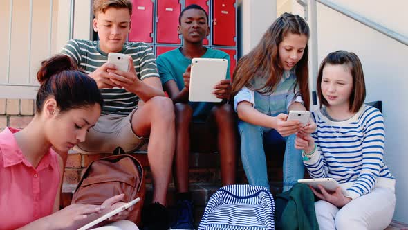 Group of smiling school friends on staircase using mobile phone and digital tablet