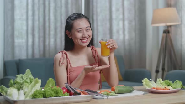 Happy Asian Woman Holding And Pointing A Glass Of Orange Juice While Having Healthy Food At Home