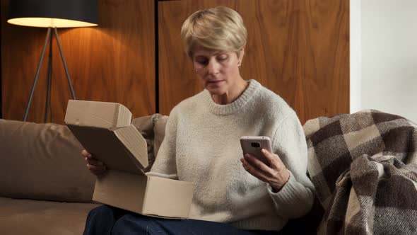 Middleaged Woman Sitting on the Sofa Opens the Box Checks the Parcel Using a Smartphone