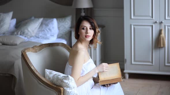 Woman in white dress sitting in armchair in vintage interior and reading a book