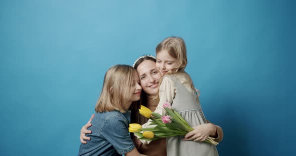 Mom Hugs Daughters and Kisses with Flowers Posing on a Blue Background