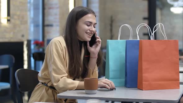 Positive Young Beautiful Woman Sitting in Cafe with Shopping Bags and Talking on the Phone