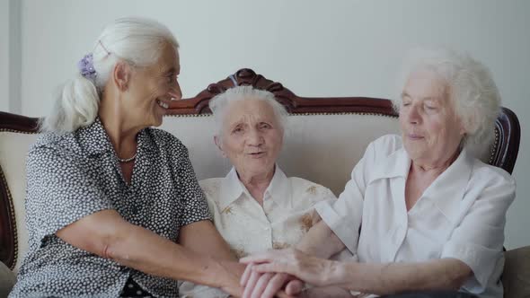 Three Cute Grandmothers Sitting on Sofa Putting Hands on Hands and Smiling