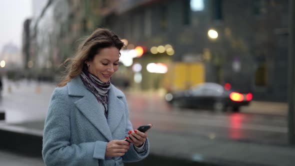 a Woman in a Light Coat and Scarf and with Bright Nails Stands at Dusk Against the Background of a