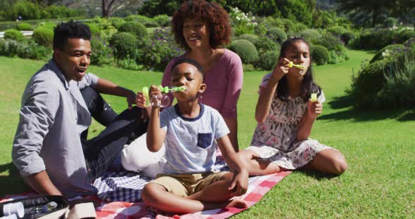 Happy african american parents, daughter and son sitting outdoors on blanket, blowing bubbles