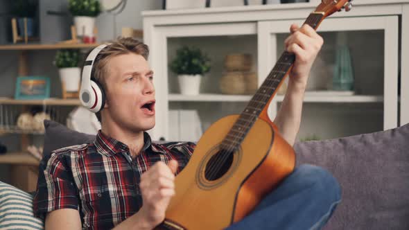 Cheerful Male Student Is Having Fun at Home Listening To Music in Headphones, Playing the Guitar