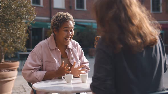 Two Diverse Female Best Friends Spending Time Together Sitting with Gift on Terrace in Cafe