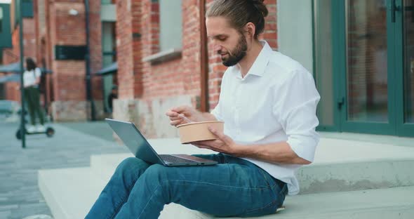 Guy Eating Takeaway Healthy Salad Outdoors and Watching Videos on Laptop, Relaxing on Stairs