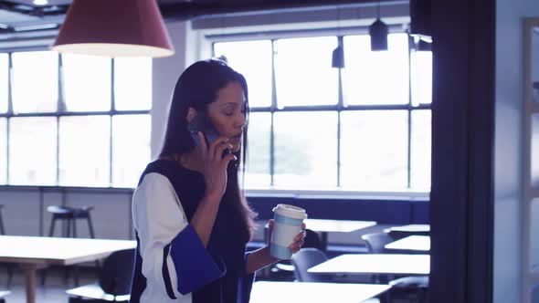 Mixed race businesswoman talking on smartphone holding coffee walking in office corridor