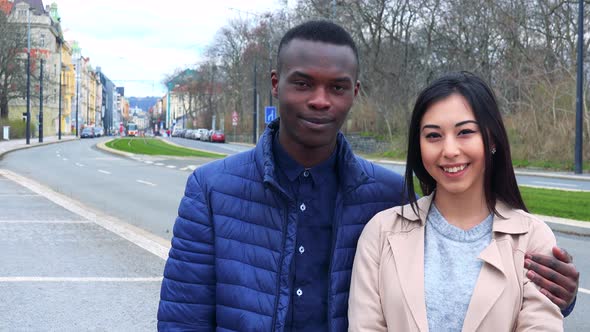 A Young Black Man and a Young Asian Woman Smile at the Camera on a Sidewalk, an Urban Area