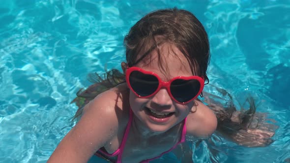 Kid Girl in Red Heart Shaped Sunglasses in Swimming Pool on Summer Sunny Day