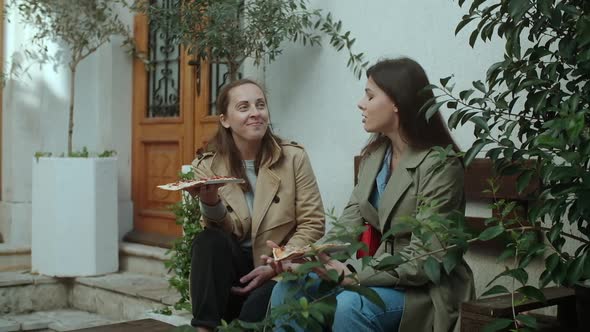 Two young women in coats eating pizza outdoors, having fun together on the terrace in spring time