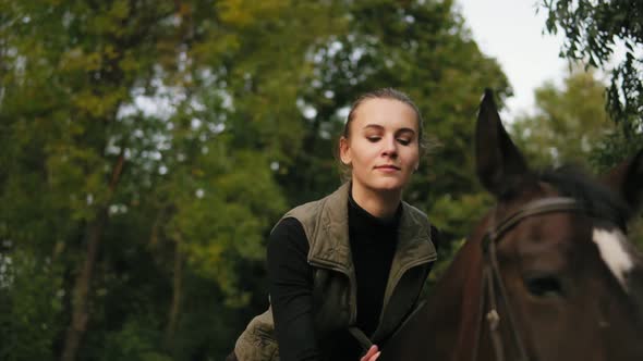 Closeup View of Young Beautiful Woman Stroking Brown Horse While Sitting in the Saddle and Putting