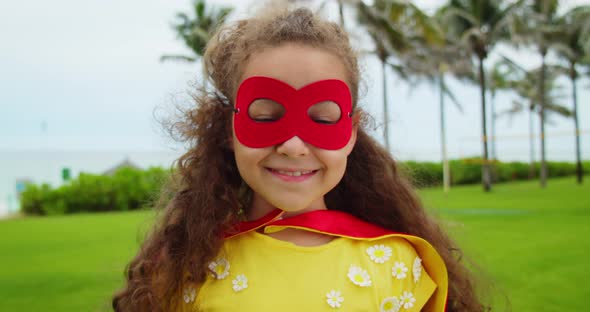 Portrait of a Cute Funny Child Girl in a Superhero Costume in a Red Cloak and a Red Mask Playing a