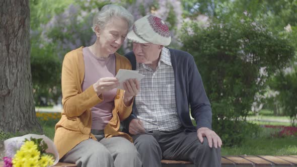 Old Woman with Bouquet of Yellow Flowers Sit in the Bench in Green Beautiful Park with an Old Man