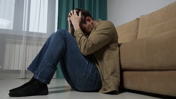 Sad Upset Man Sits on Floor Near Sofa in Living Room