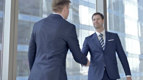 Two Middle Aged Businessmen Shaking Hand and Passing Money Against Office Window