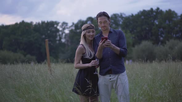 Positive Couple Standing on Windy Summer Meadow in the Evening and Using Smartphone. Portrait of