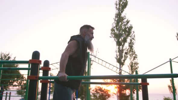 Fit Handsome Middle Aged Man with Long Gray Beard Doing Push Ups on Parallel Bars on Sports Ground