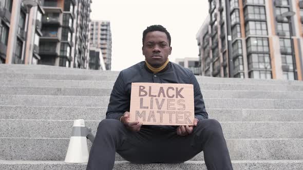 Young Man Sitting on the Stairs and Holding Carton Placard with Black Lives Matter Writing