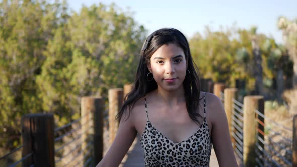 Cute hispanic woman walking in slow motion along a wooden bridge outdoors in sunlight smiling and la