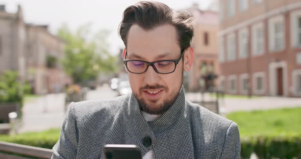 Young Man Wearing Glasses with Mustaches and a Beard is Sitting on a Bench in the Square Reading