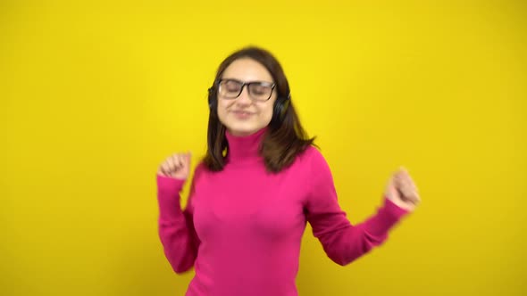 Young Woman Dancing with Headphones on a Yellow Background. A Girl in a Pink Turtleneck and Glasses