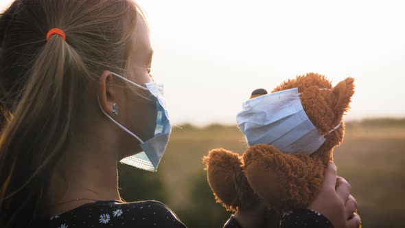 Sad Little Girl and Teddy Bear with Surgical Mask at Sunset. A Child Holding Her Teddy Bear During