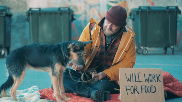 A Homeless Man is Feeding His Dog While Sitting on the Ground